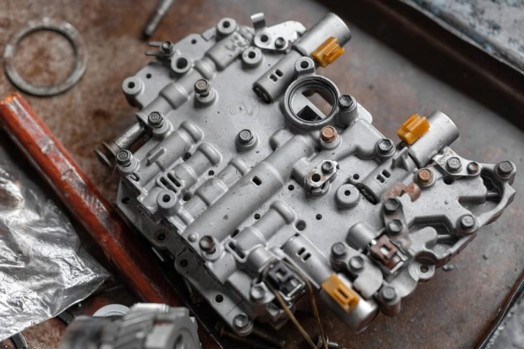 A top-down, detailed view of an automatic transmission valve body removed from a vehicle and resting on a workbench, showing its complex hydraulic passages and electronic solenoids.